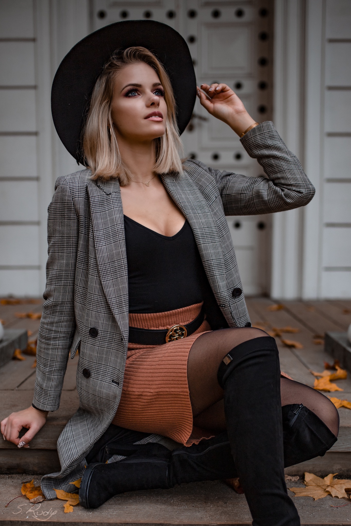 Kobieta w kraciastej marynarce, czarnym kapeluszu i kozakach za kolano pozuje siedząc na drewnianym ganku ozdobionym opadłymi liśćmi.