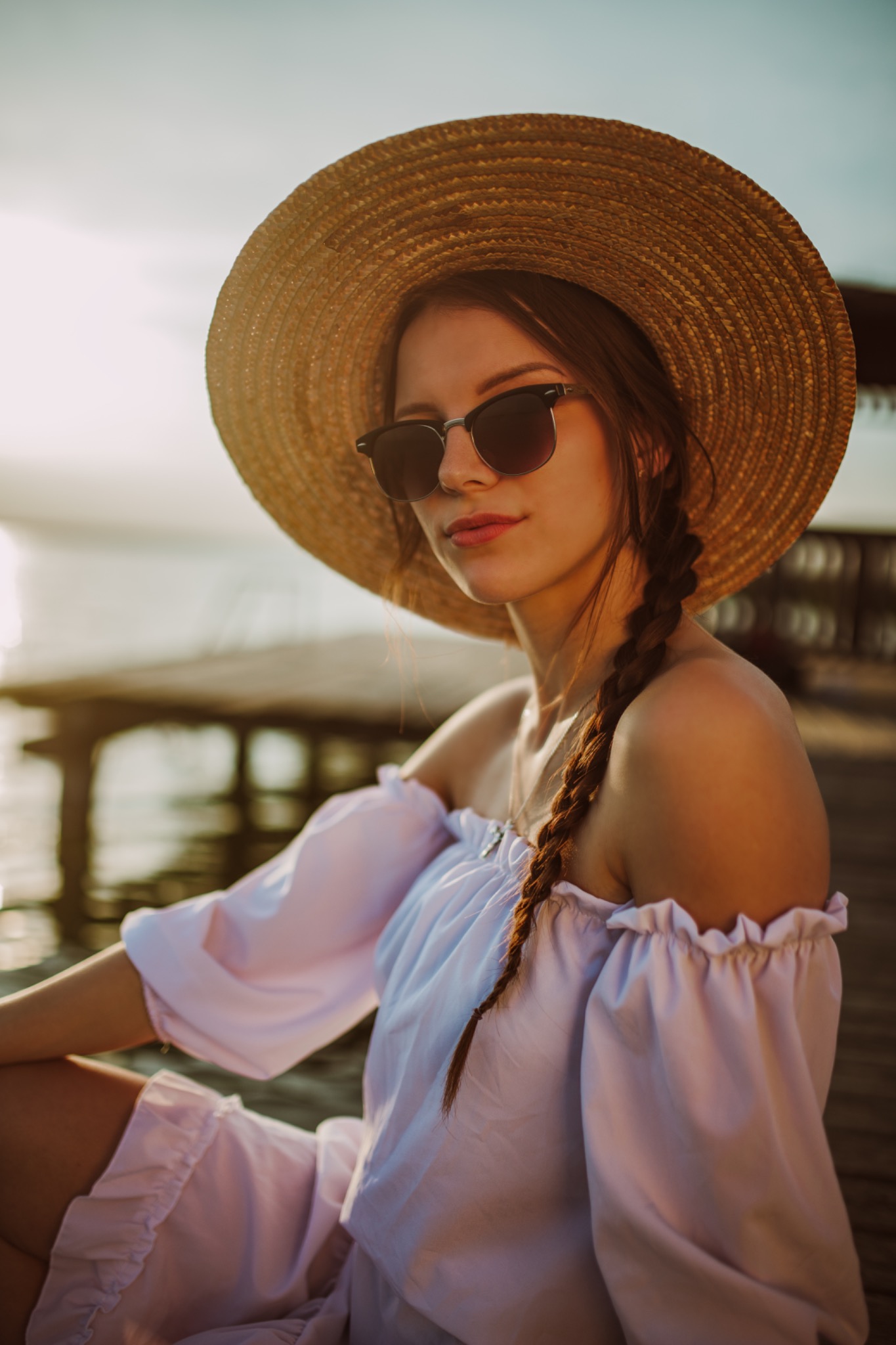 Portret młodej kobiety w słomkowym kapeluszu i okularach przeciwsłonecznych, siedzącej na drewnianym pomoście nad wodą, z warkoczem i bluzką z odkrytymi ramionami.