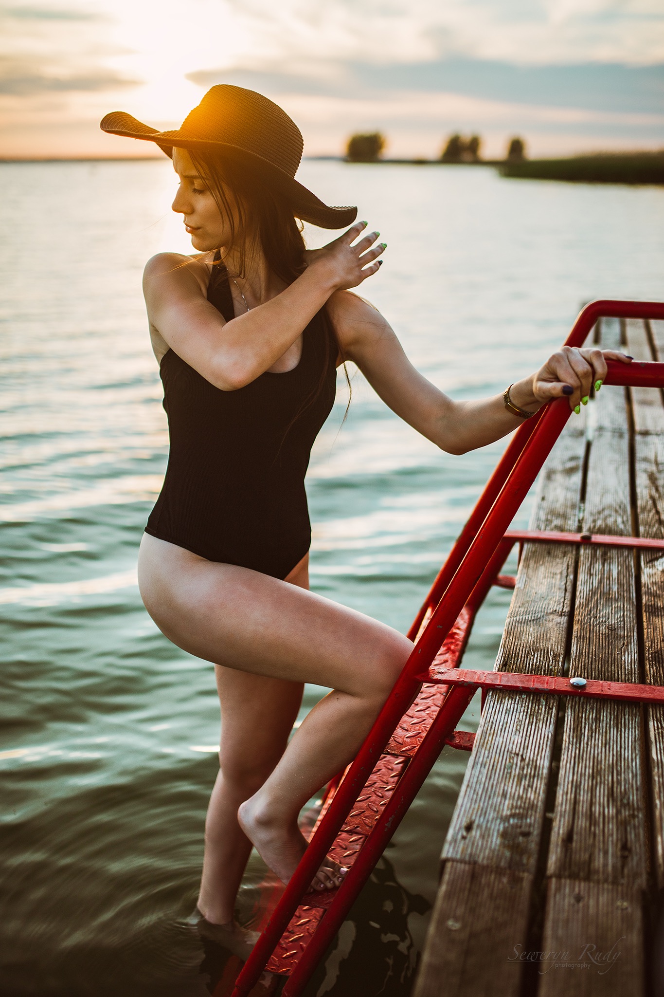Kobieta w czarnym stroju kąpielowym i kapeluszu wchodzi po czerwonej drabince do wody, o zachodzie słońca.
