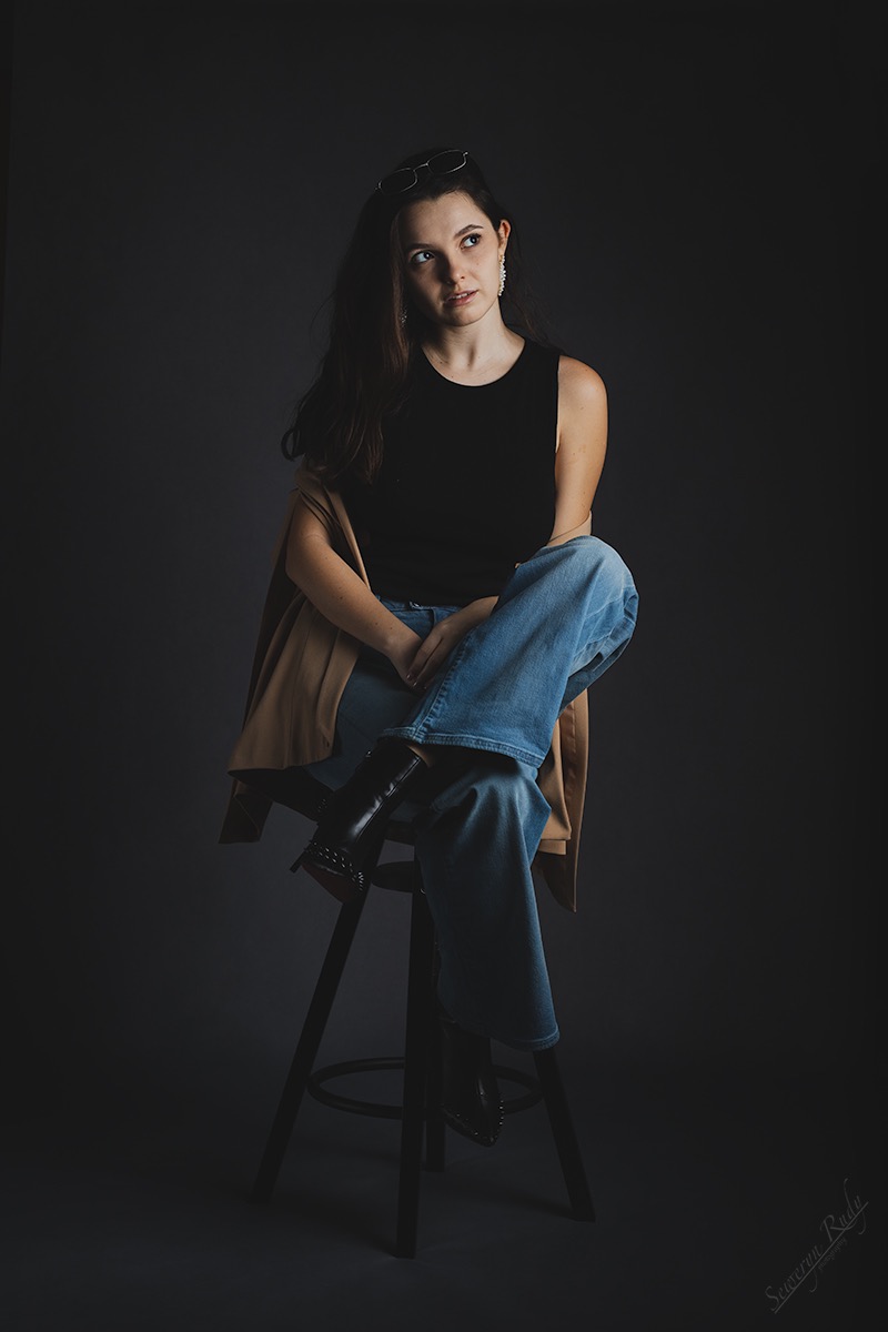 Portret kobiety siedzącej na stołku, ubrana w dżinsy i czarny top, z marynarką zarzuconą na ramiona, okulary na głowie i błyszczące kolczyki, spoglądającej w lewo.