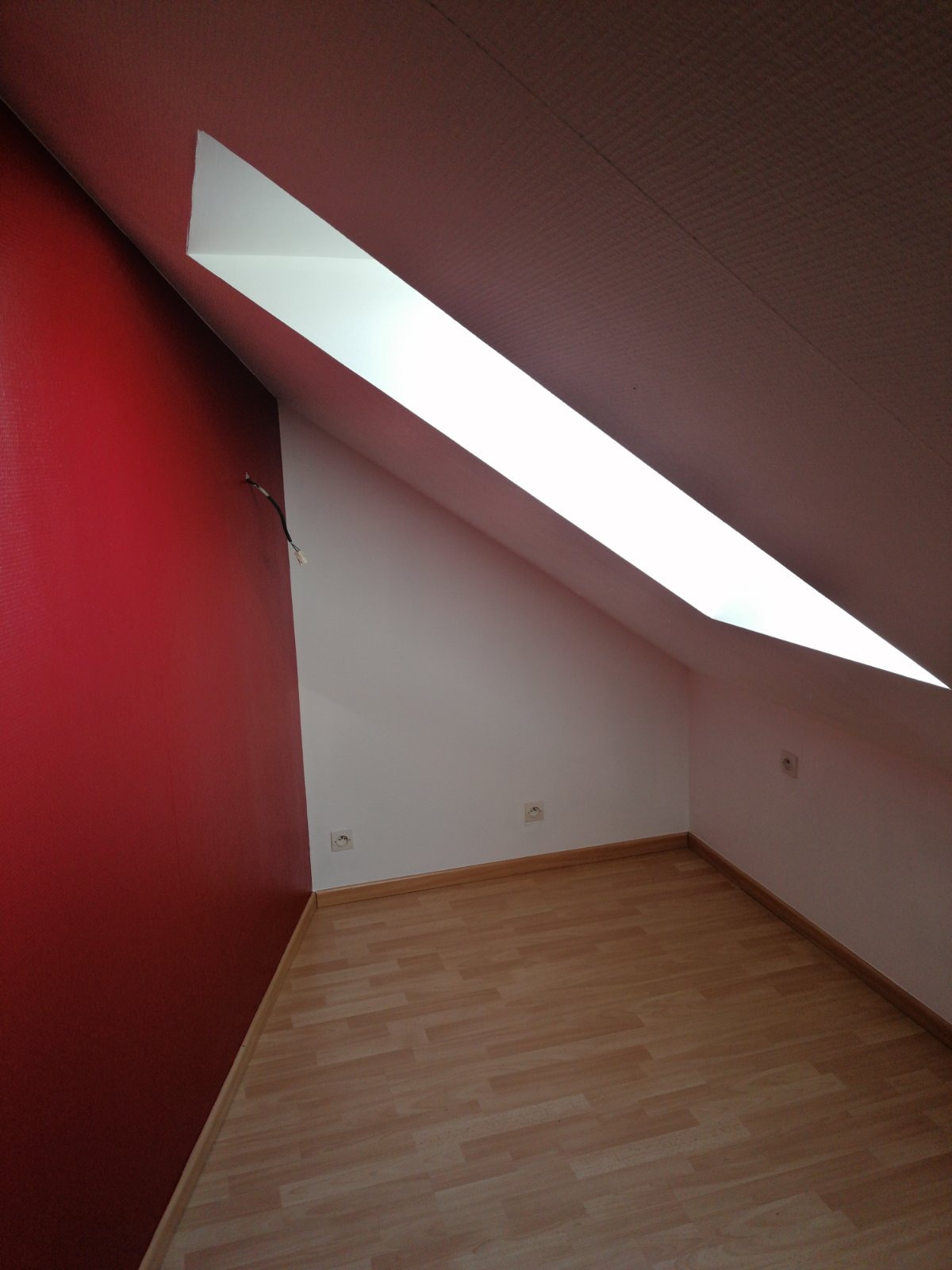 Poddasze po remoncie: czerwona ściana kontrastuje z białymi ścianami i podłogą z paneli, widoczne gniazdka elektryczne i wystający kabel.