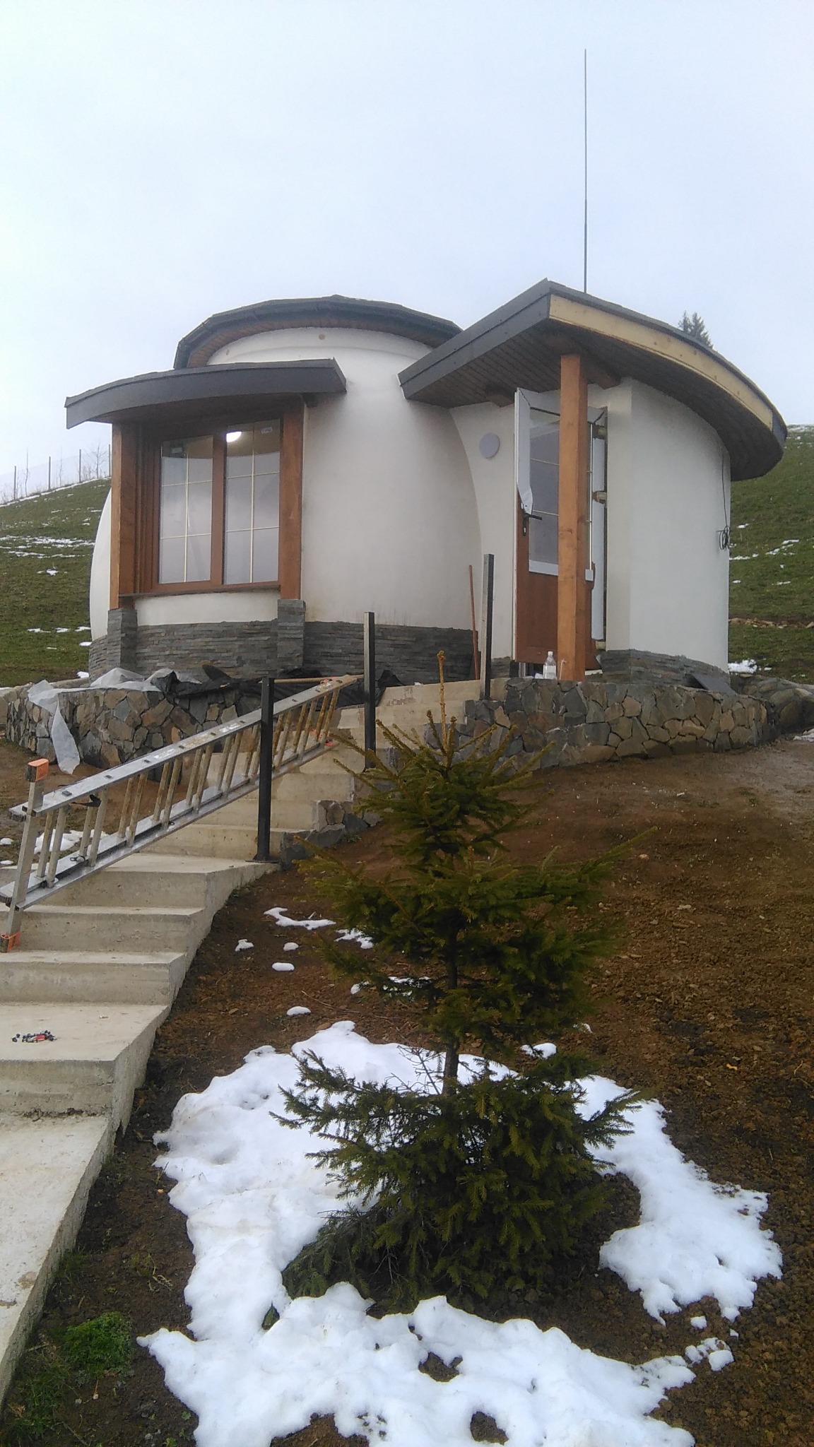 Nietypowy, okrągły dom w trakcie budowy na wzgórzu, z widocznymi schodami wejściowymi, drabina oparta o schody i fragmentami śniegu na ziemi.