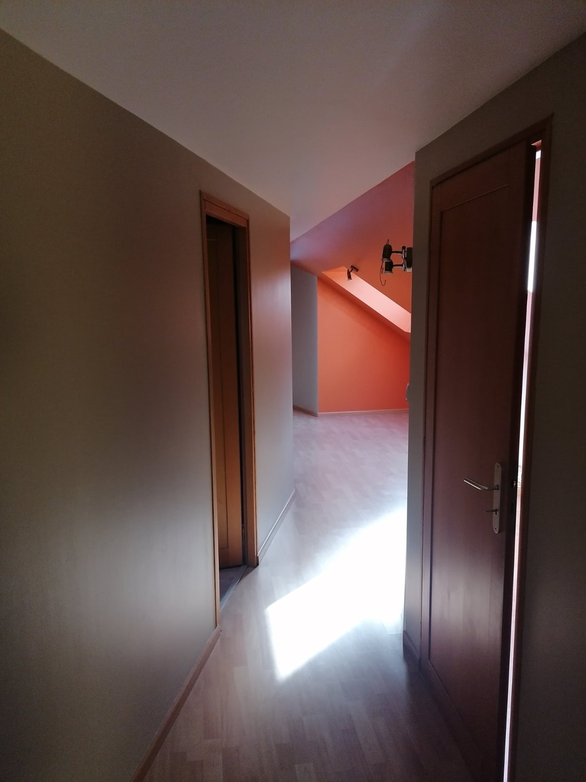 Korytarz po remoncie z parkietem, świeżo pomalowanymi ścianami w odcieniach beżu i pomarańczy, widoczny przez otwarte drzwi, z lampami sufitowymi. W tle widoczne poddasze.