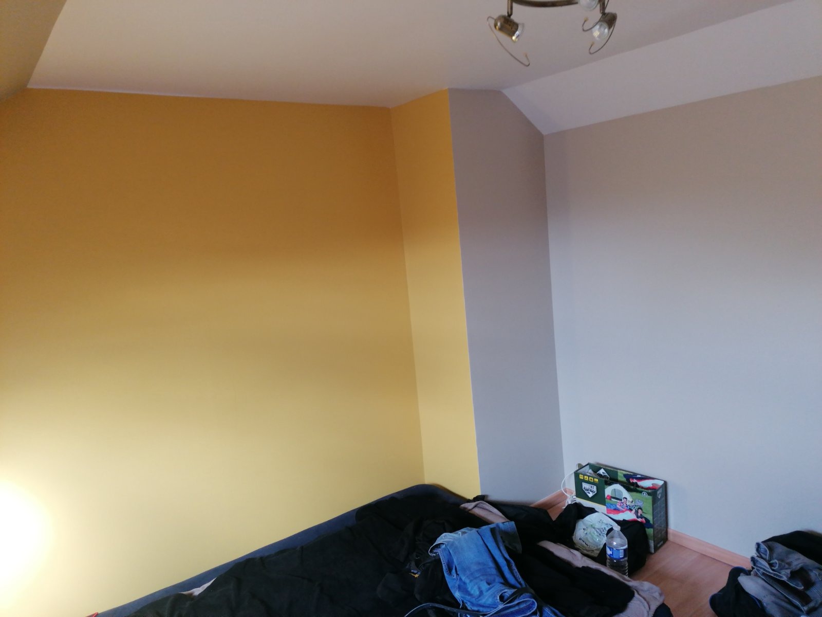 Pokój po malowaniu: żółta ściana kontrastuje z szarą, widoczne ubrania i karton na podłodze.