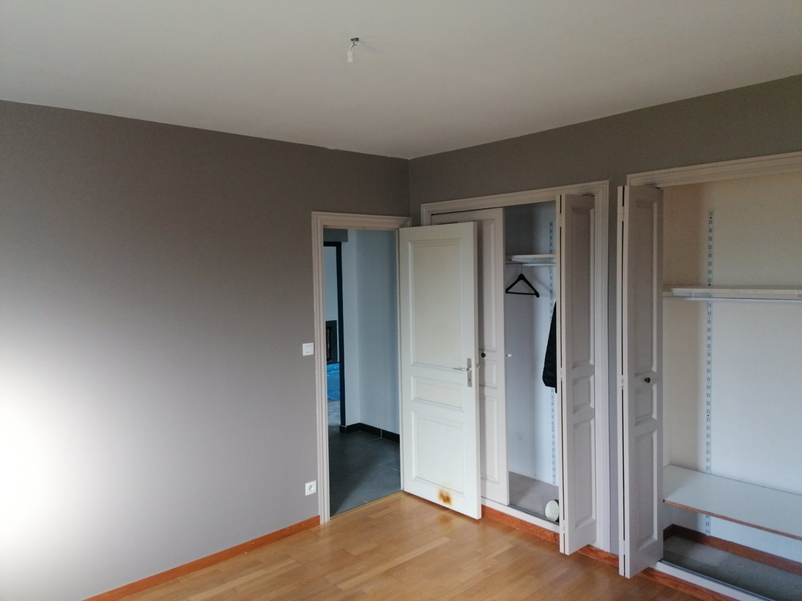 Pokój po malowaniu ścian na szaro, z otwartymi drzwiami do innego pomieszczenia i szafami w zabudowie z widocznymi półkami i wieszakami.