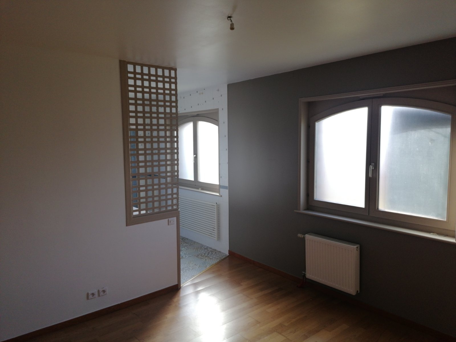 Pusty pokój po remoncie z drewnianą podłogą, szarą ścianą z oknem, białą ścianą z ażurową ścianką działową i widocznym fragmentem innego pomieszczenia z białą ścianą i oknem łukowym.