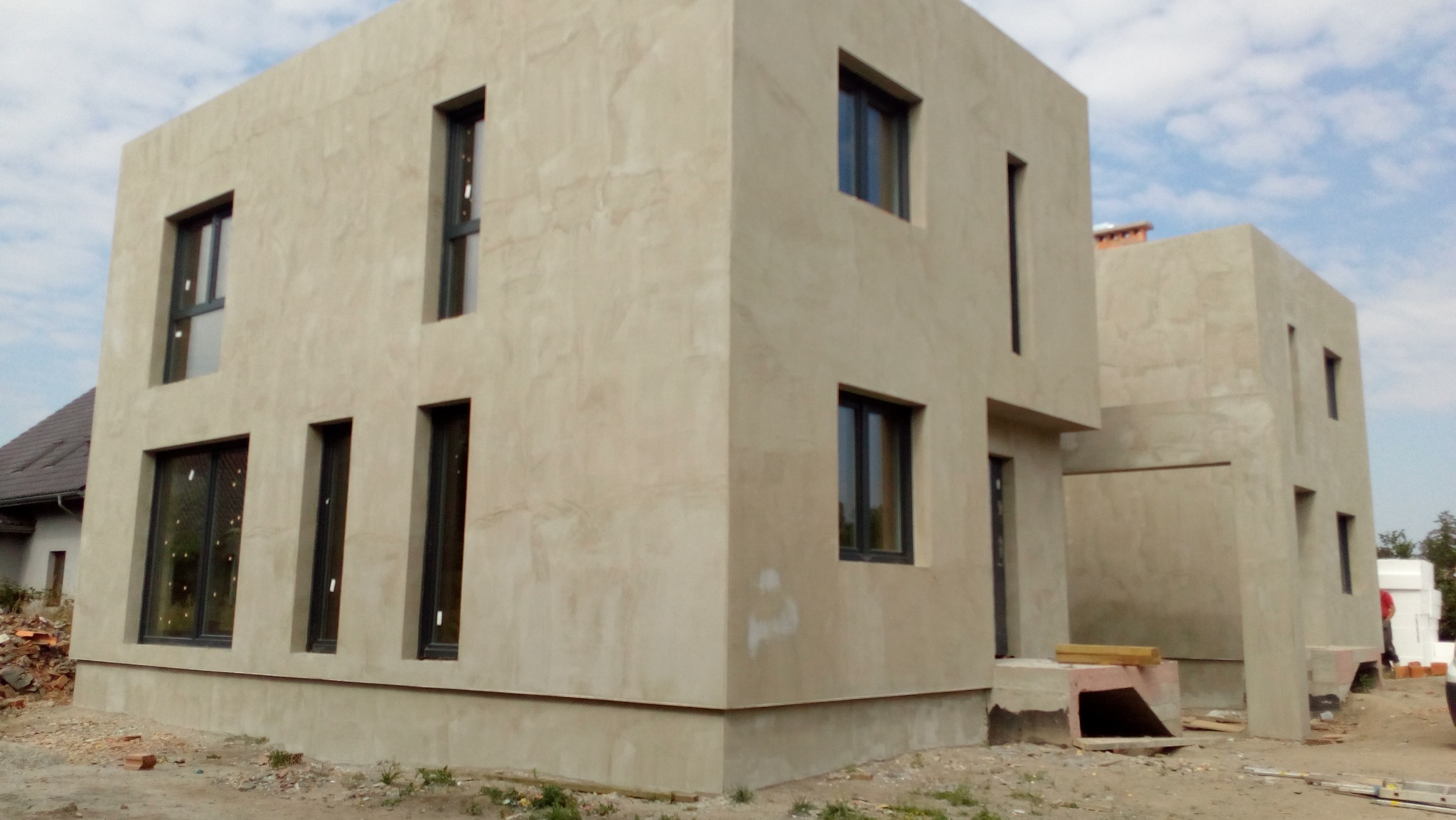 Surowy budynek w stylu minimalistycznym z beżową elewacją i ciemnymi oknami, widoczne ślady po ocieplaniu, teren wokół niezagospodarowany.