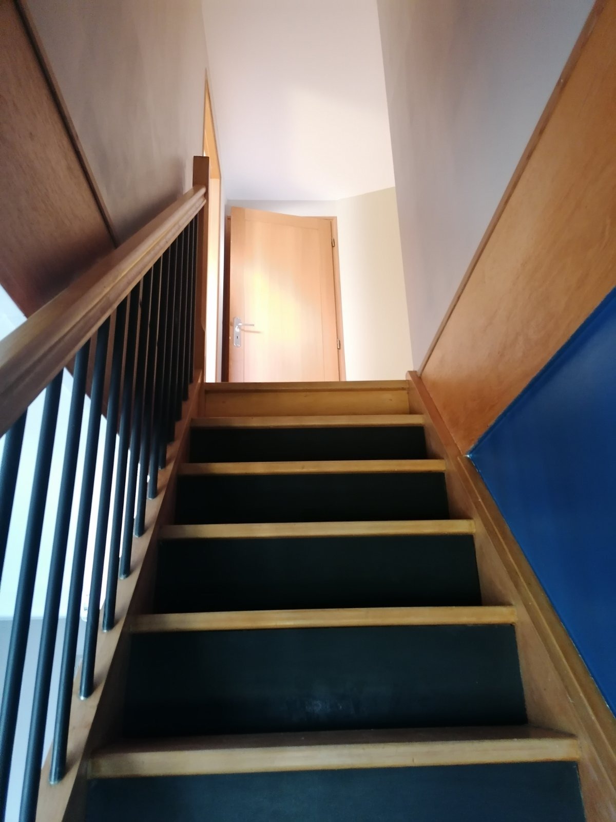 Drewniane schody z czarnymi stopniami i metalową balustradą prowadzące do jasnych drzwi.