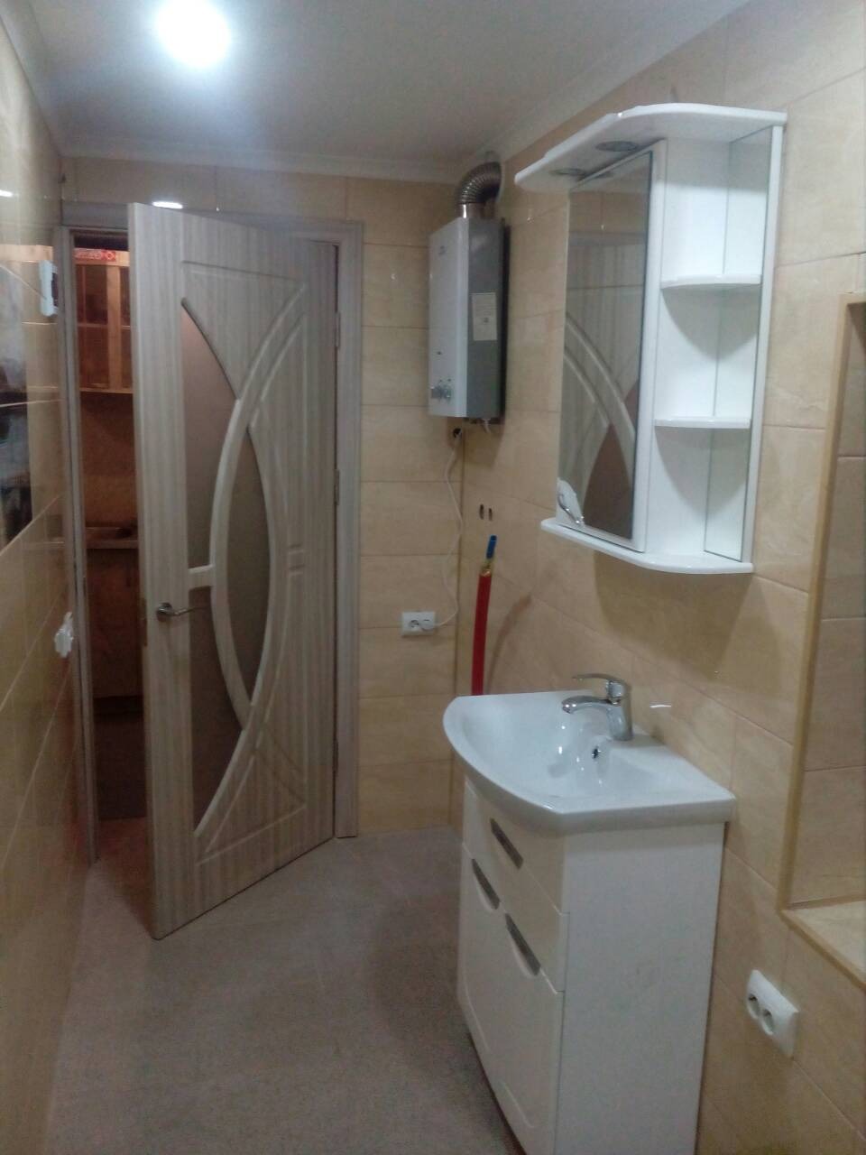 Wykończona łazienka z beżowymi płytkami, białą szafką z umywalką, lustrzaną szafką wiszącą i grzejnikiem gazowym na ścianie, widoczne otwarte drzwi do innego pomieszczenia.