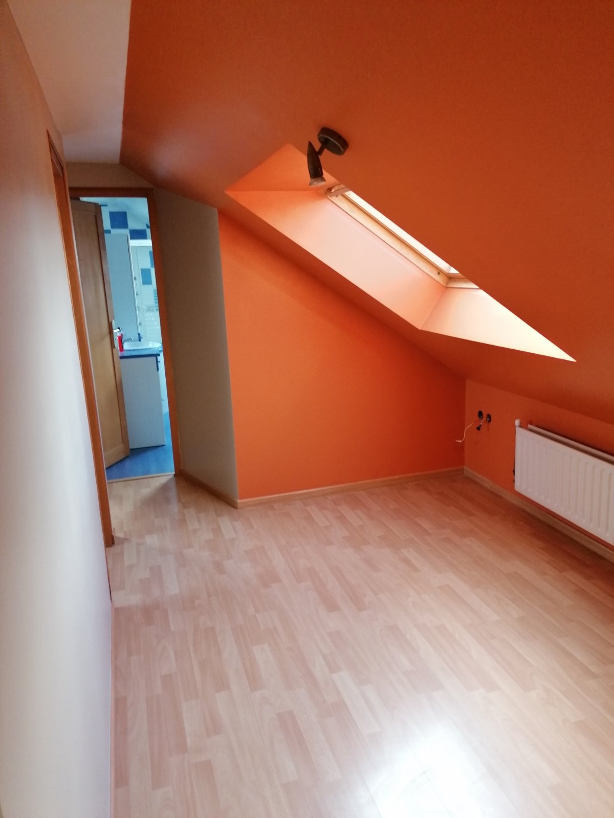 Poddasze po remoncie: pomarańczowe ściany, jasne panele podłogowe, okno dachowe z lampą, widok na sąsiednie pomieszczenie z niebieskimi akcentami.