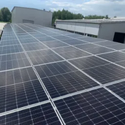 Rozległa instalacja paneli słonecznych na dachu budynku przemysłowego, widok z góry, z budynkami w tle i fragmentem zieleni po lewej stronie.