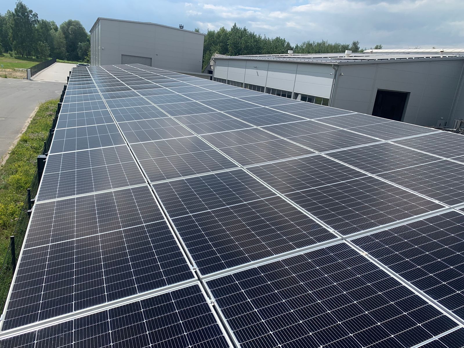 Rozległa instalacja paneli słonecznych na dachu budynku przemysłowego, widok z góry, z budynkami w tle i fragmentem zieleni po lewej stronie.