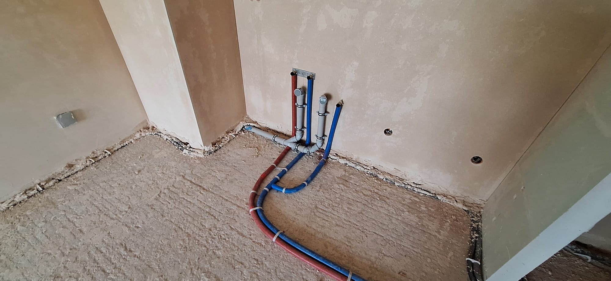 Fragment wnętrza w trakcie prac instalacyjnych: widoczne rury miedziane i plastikowe, mocowania na ścianie i bruzdy w podłodze przygotowane pod instalacje.