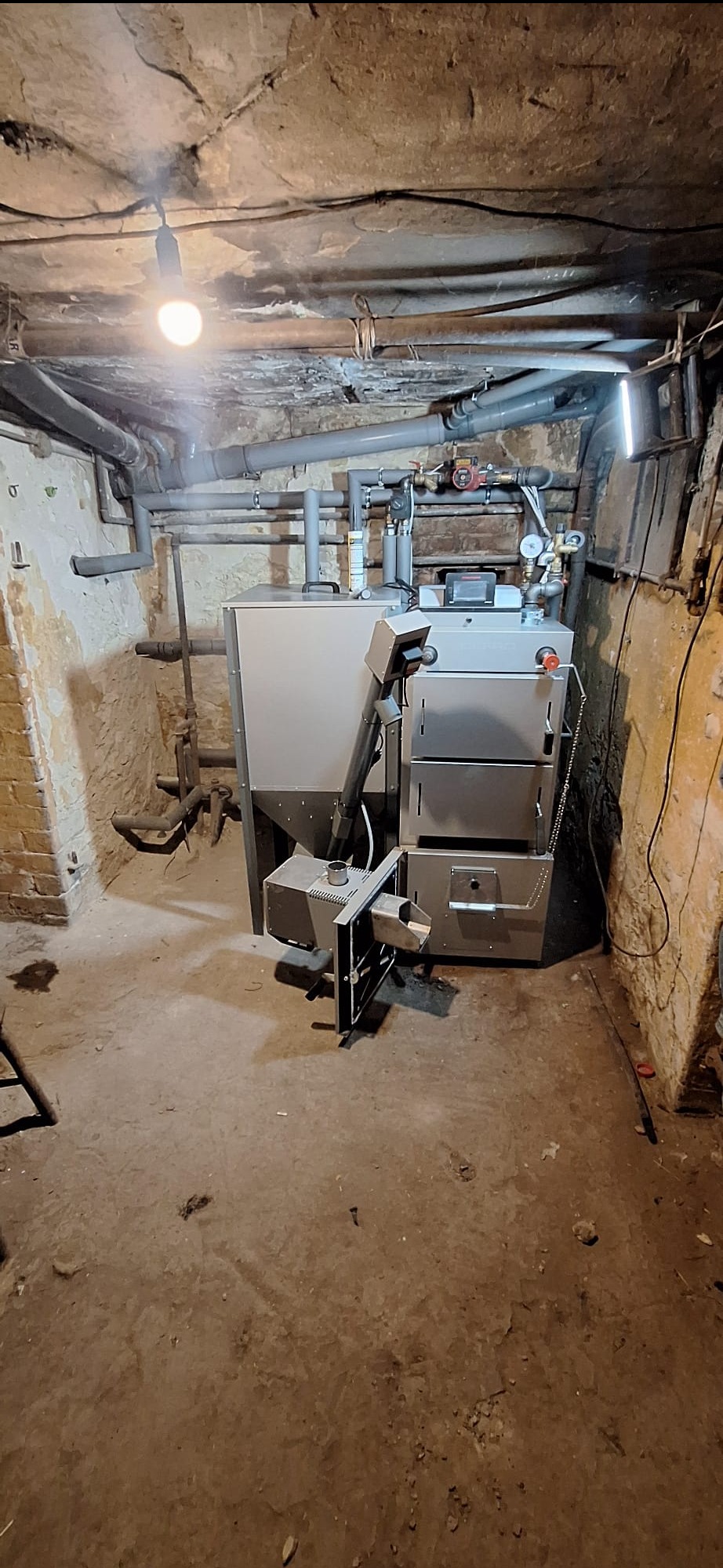 Nowoczesny piec na paliwo stałe zainstalowany w piwnicy o surowych ścianach. Widoczne rury instalacji grzewczej i oświetlenie żarówką.