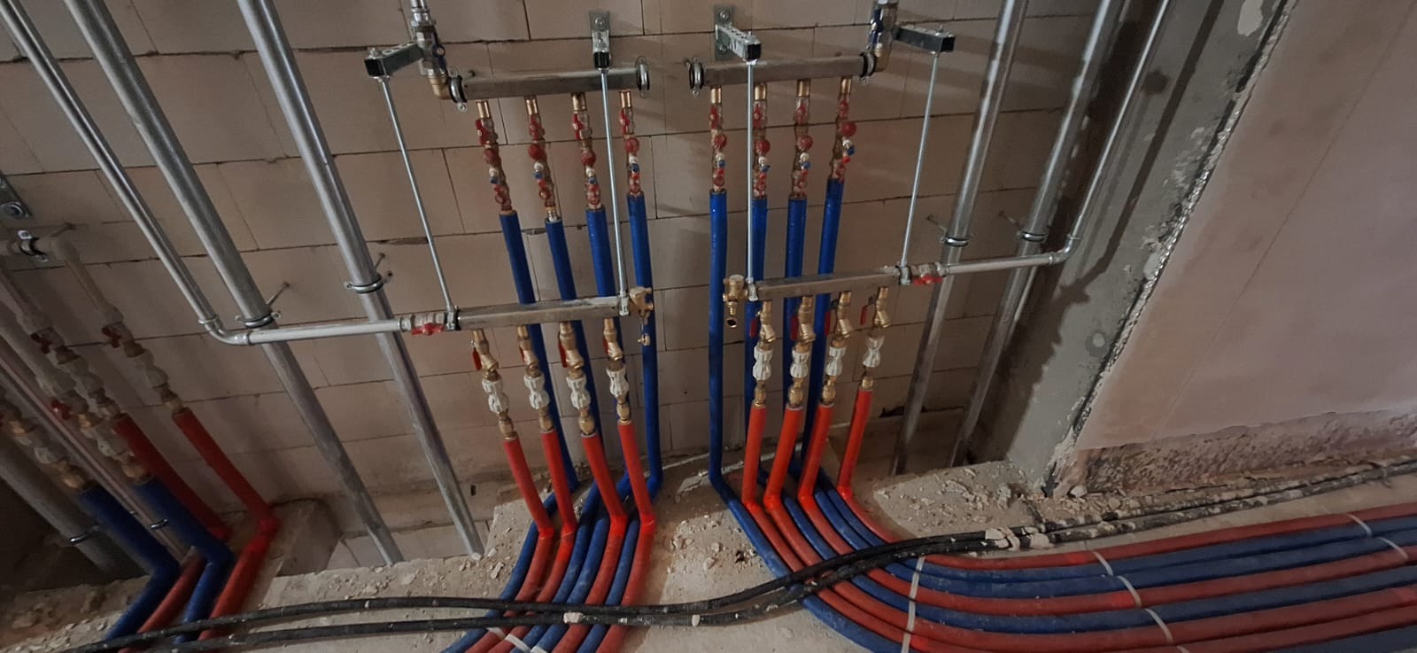 Rozdzielacz ogrzewania podłogowego z rurami PEX w kolorze niebieskim i czerwonym, zamontowany na ścianie z bloczków, widok z dołu podczas instalacji.