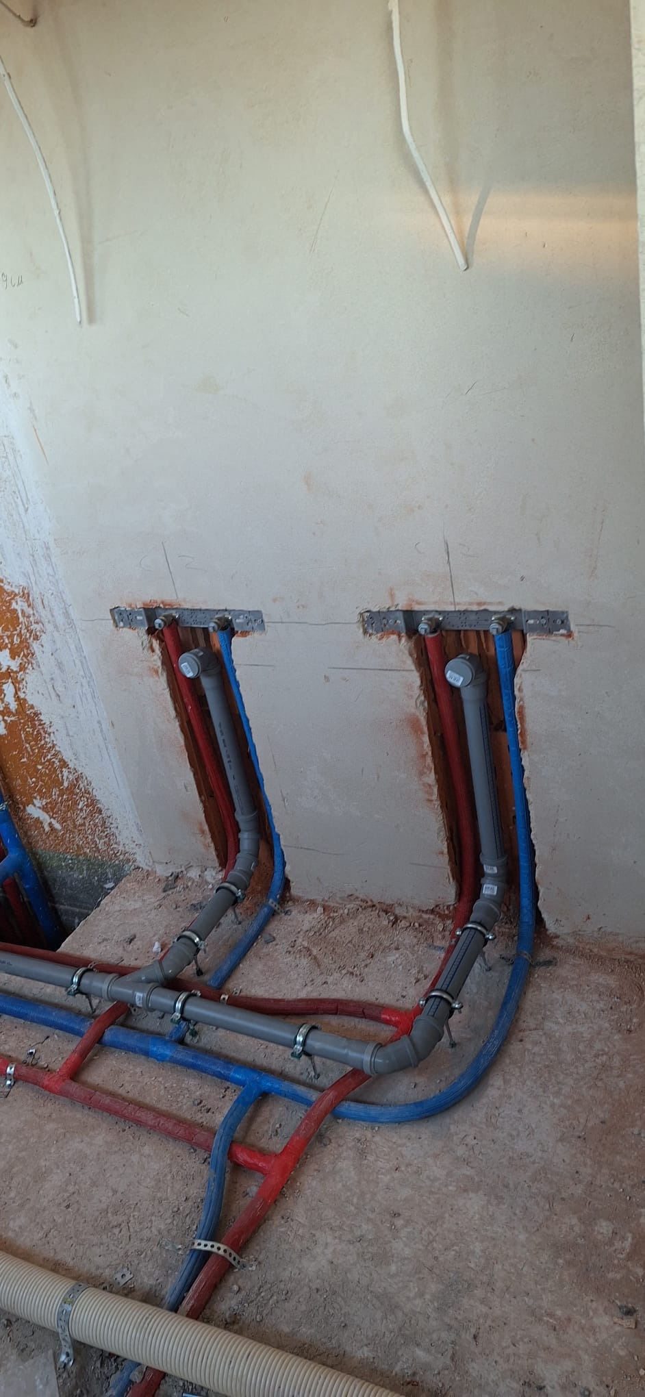 Montaż instalacji wodnej i kanalizacyjnej w bruzdach ściennych, rury z tworzywa w kolorach czerwonym i niebieskim, szare rury kanalizacyjne, etap prac instalacyjnych.