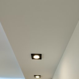 Remonty wykończenia-ocieplenia - Biały sufit z zamontowanymi, kwadratowymi lampami LED w czarnych oprawach, ułożonymi w rzędzie, z widocznym fragmentem ściany po prawej stronie.