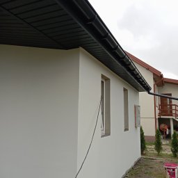 Remonty wykończenia-ocieplenia - Świeżo pomalowana, biała ściana domu jednorodzinnego z czarnym orynnowaniem i podbitką dachową; widoczna puszka farby Ceresit CT48.