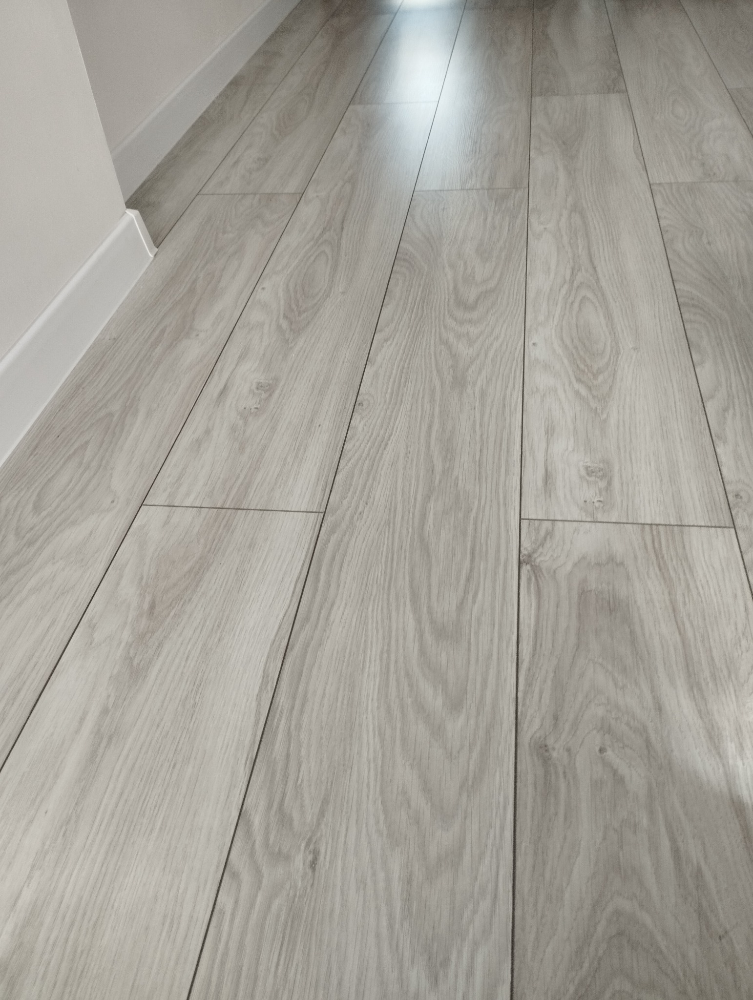 Jasne, drewnopodobne panele podłogowe ułożone wzdłuż ściany z białą listwą przypodłogową w Łyse. Widoczne delikatne słoje i subtelne przejścia tonalne na powierzchni paneli.