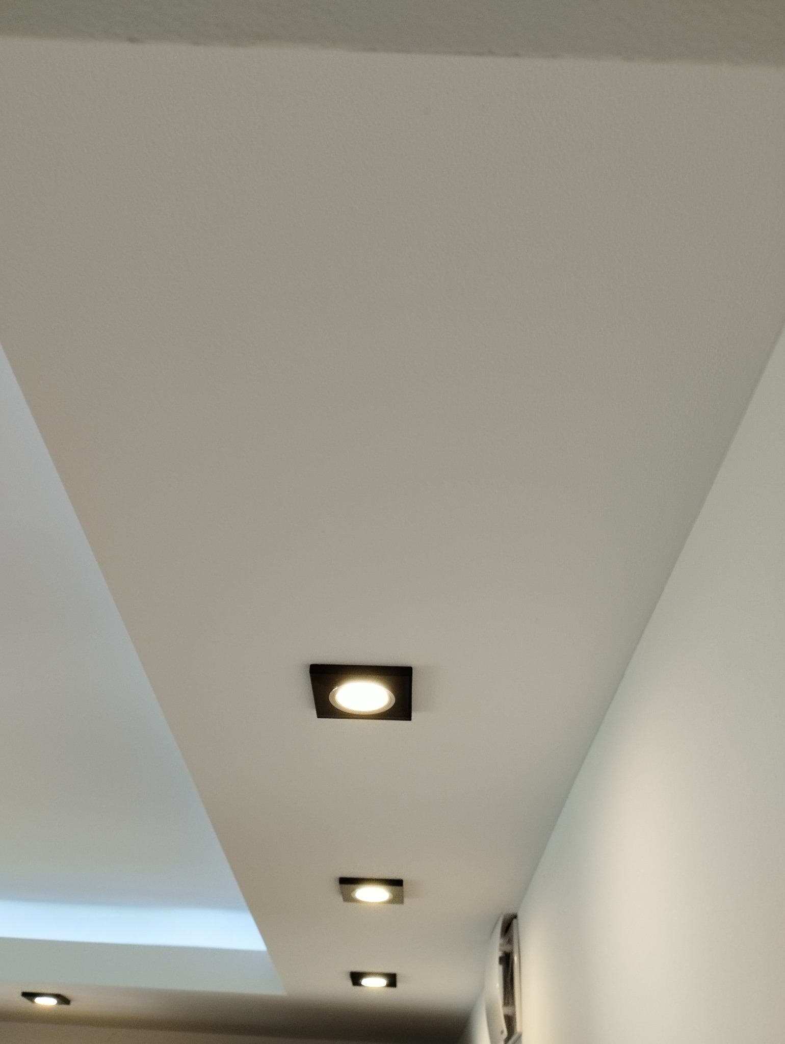 Biały sufit z zamontowanymi, kwadratowymi lampami LED w czarnych oprawach, ułożonymi w rzędzie, z widocznym fragmentem ściany po prawej stronie.