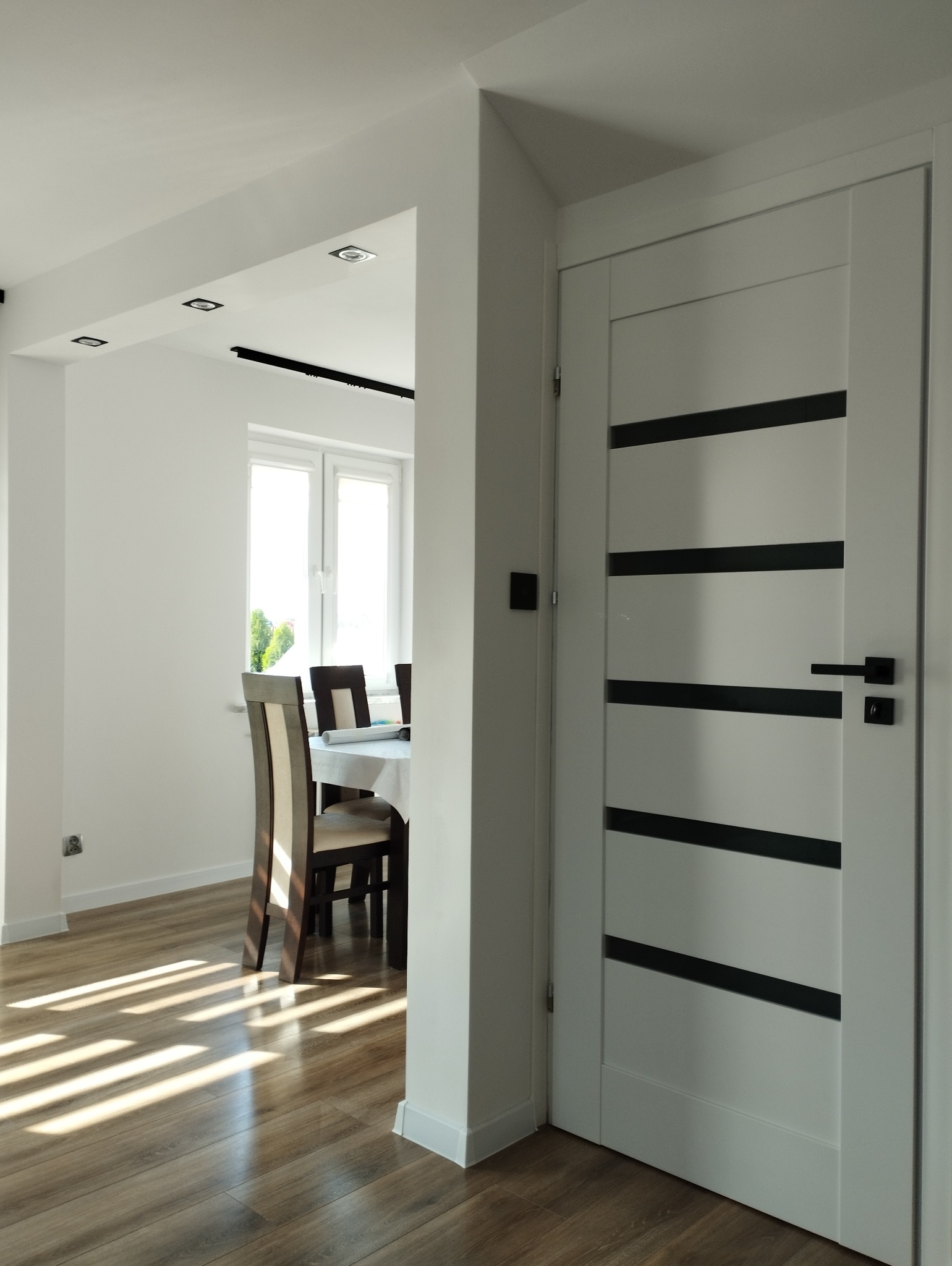 Nowoczesne wnętrze z białą ścianą, drewnianą podłogą i drzwiami z czarnymi poziomymi paskami, widok na jadalnię z białym obrusem i krzesłami.