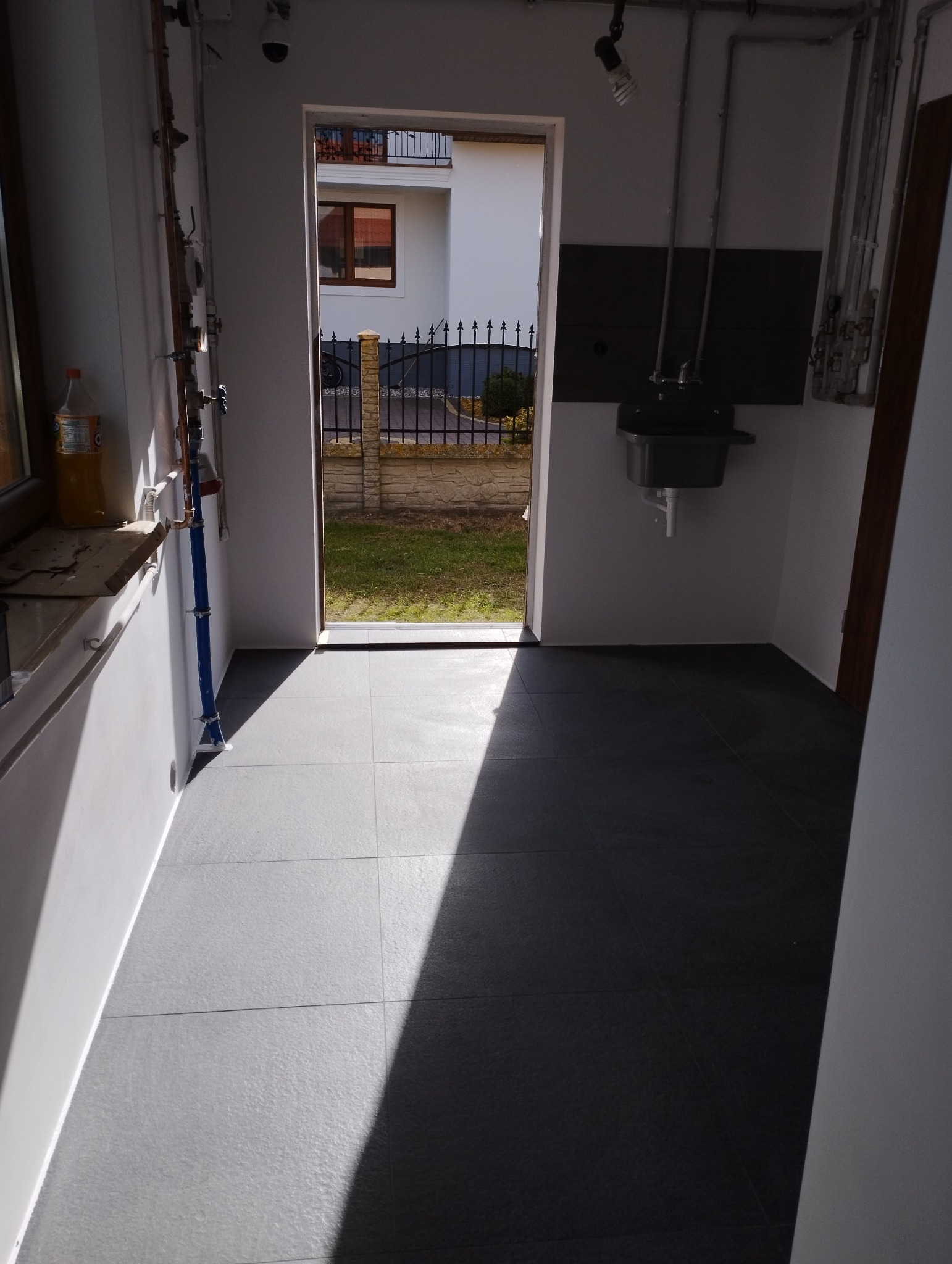 Wykończone pomieszczenie gospodarcze z ciemną, matową posadzką z płytek, umywalką na ścianie i widokiem na ogród przez otwarte drzwi, oświetlone silnym promieniem światła słonecznego.