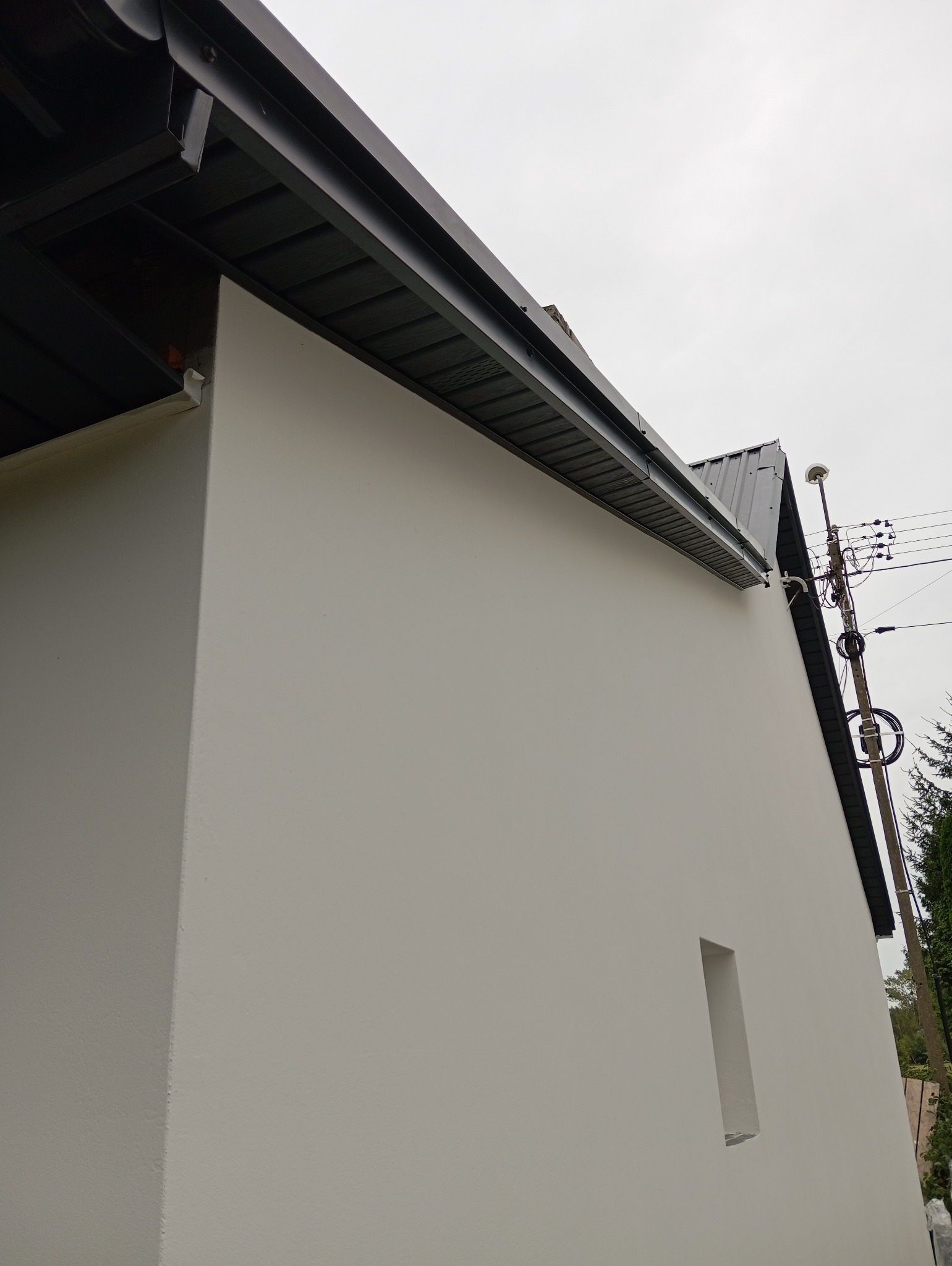 Ocieplona i pomalowana elewacja budynku z widocznym fragmentem dachu, rynny i słupa energetycznego w tle.