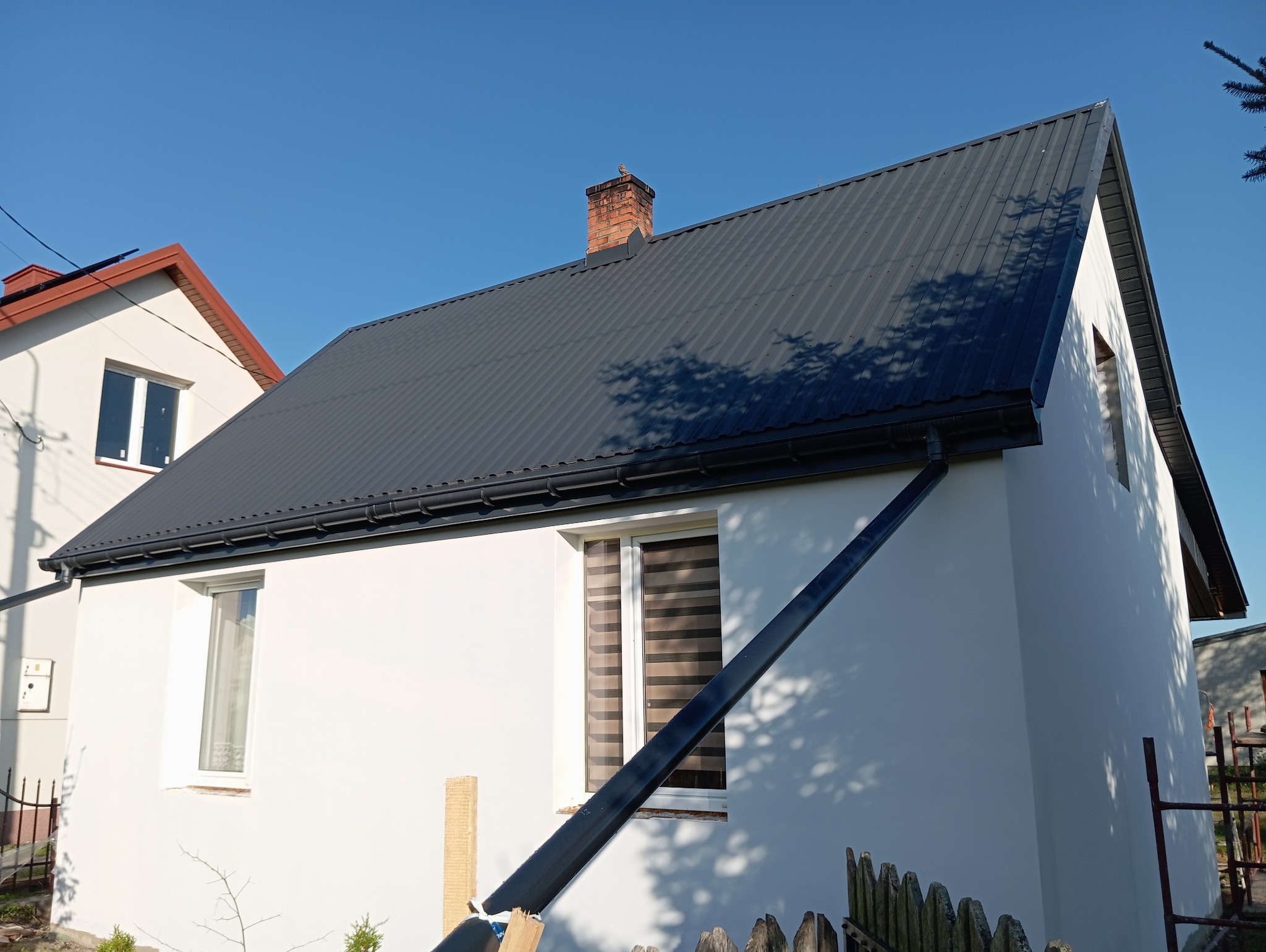 Odnowiona elewacja domu z nowym, grafitowym dachem z blachy trapezowej i świeżo pomalowanymi ścianami na biało, widoczne rynny i komin z cegły.