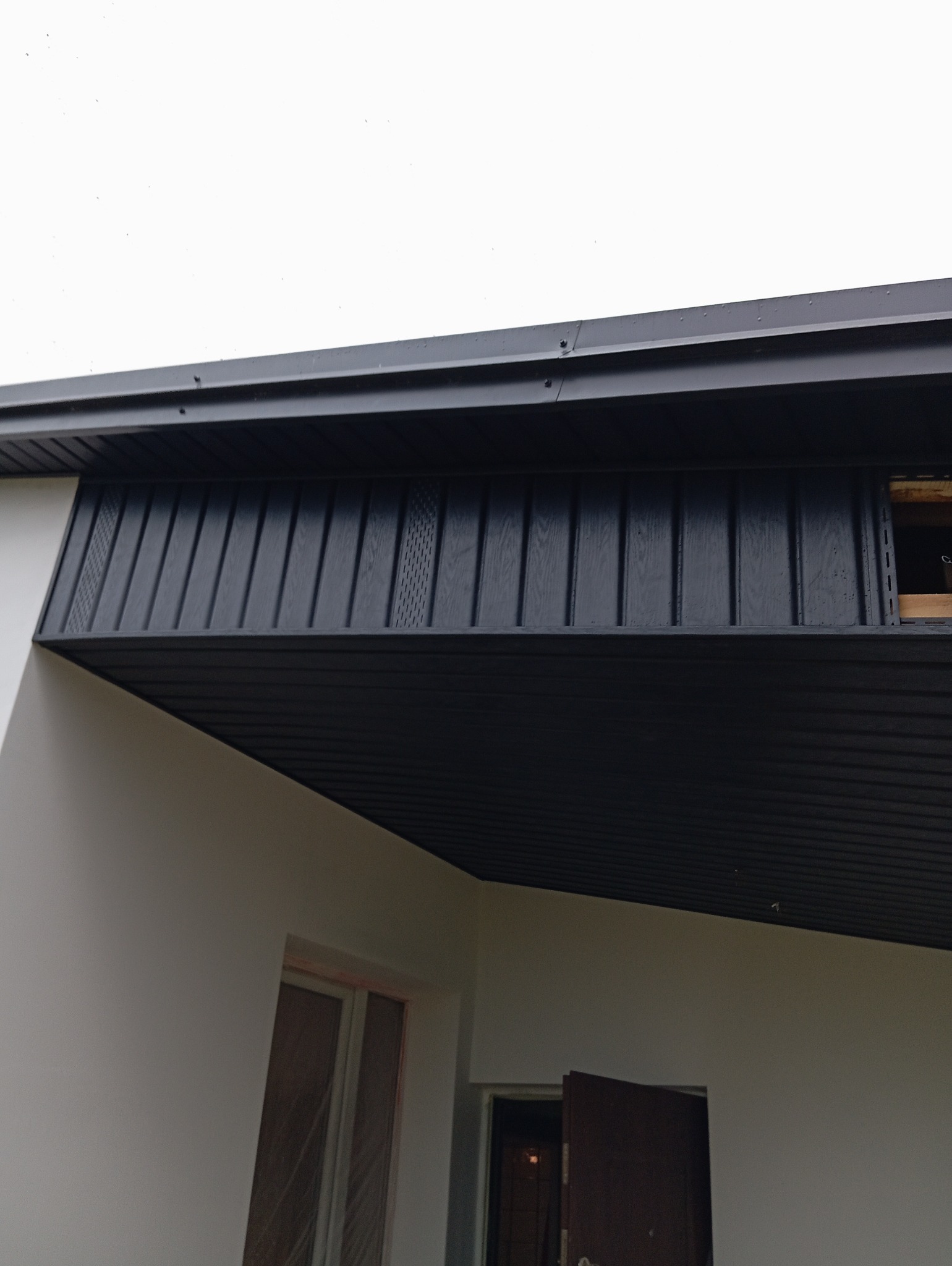 Ciemna podbitka dachowa z perforowanymi panelami wentylacyjnymi, zamontowana na elewacji domu z otwartymi drzwiami wejściowymi.