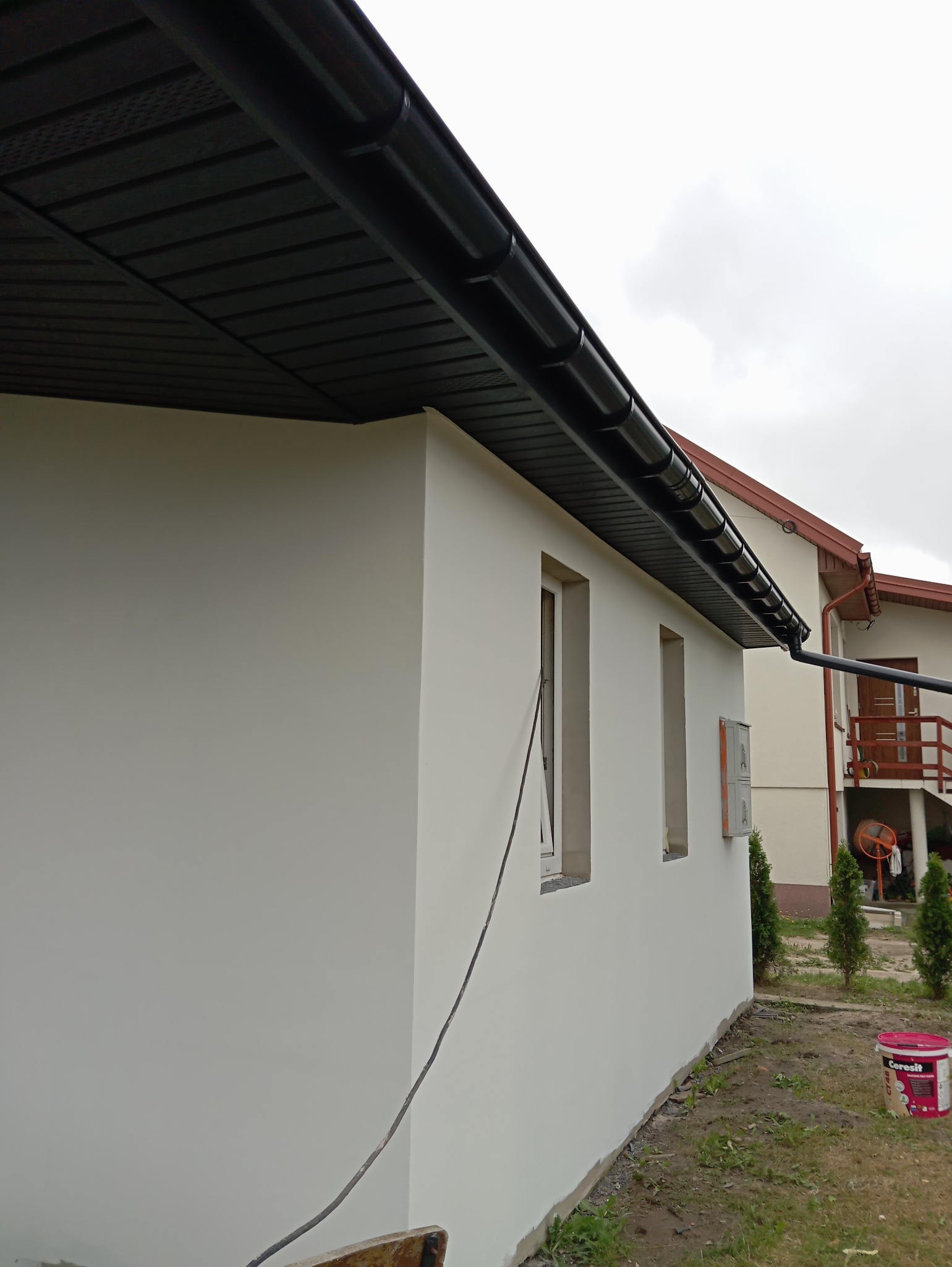 Świeżo pomalowana, biała ściana domu jednorodzinnego z czarnym orynnowaniem i podbitką dachową; widoczna puszka farby Ceresit CT48.