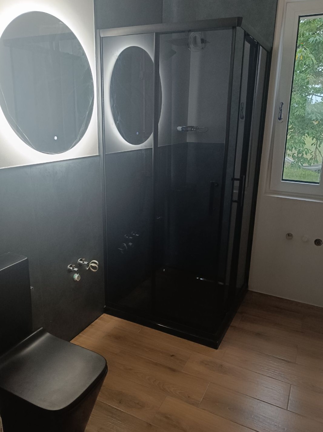 Nowoczesna łazienka z czarną kabiną prysznicową, podświetlanymi okrągłymi lustrami i drewnopodobną podłogą; widoczna czarna toaleta i fragment okna z widokiem na zieleń.