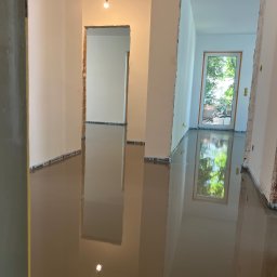 MAT BUD - Wnętrze w trakcie wylewania samopoziomującej wylewki. Widoczne przygotowane ściany z widocznymi śladami po poprzednich pracach i narzędzie do rozprowadzania wylewki.