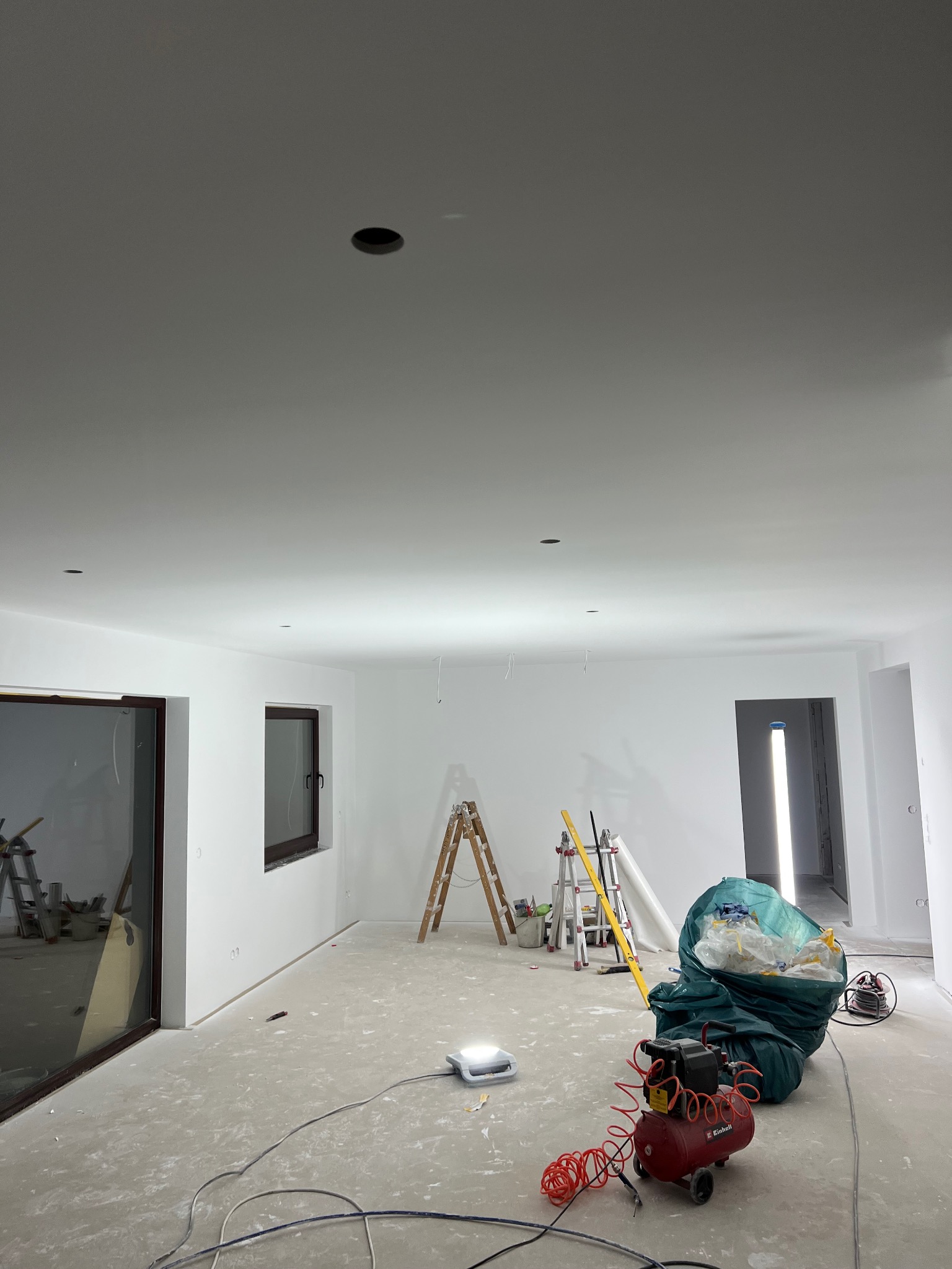 Wnętrze po szpachlowaniu ścian i sufitu, widoczne narzędzia: drabiny, kompresor, worki z odpadami. Przygotowanie do malowania.