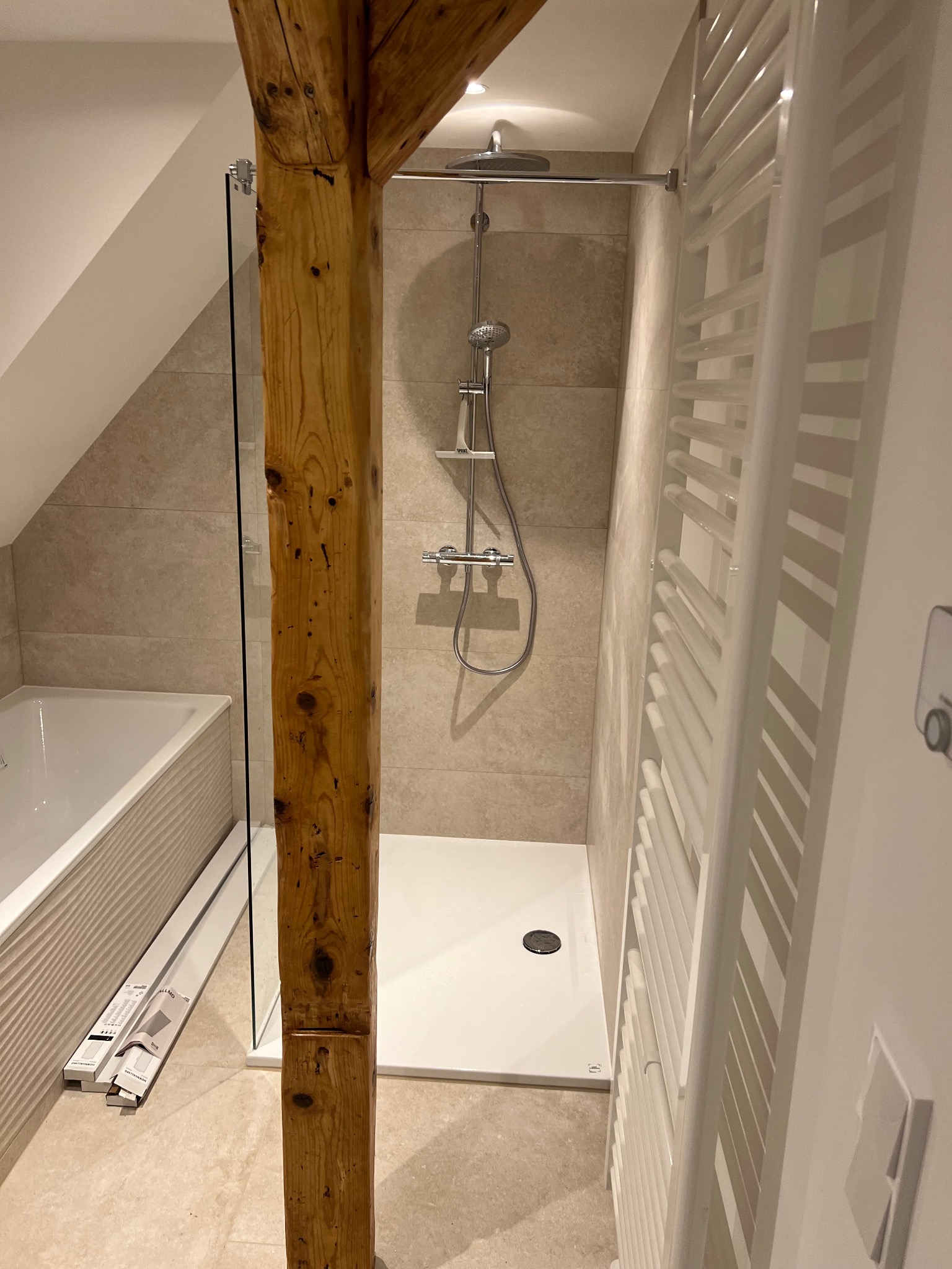 Wykończona łazienka na poddaszu z kabiną prysznicową typu walk-in, drewnianą belką konstrukcyjną i grzejnikiem drabinkowym. Beżowe płytki i biała armatura.