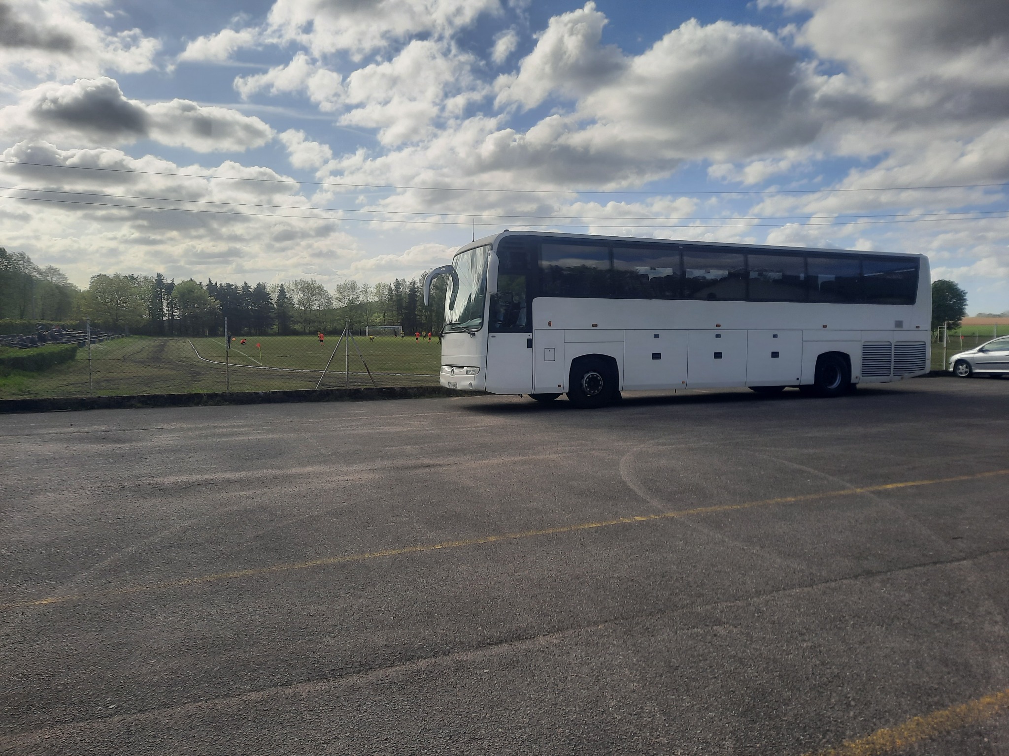 Biały autokar zaparkowany na parkingu z widokiem na boisko sportowe i pochmurne niebo.