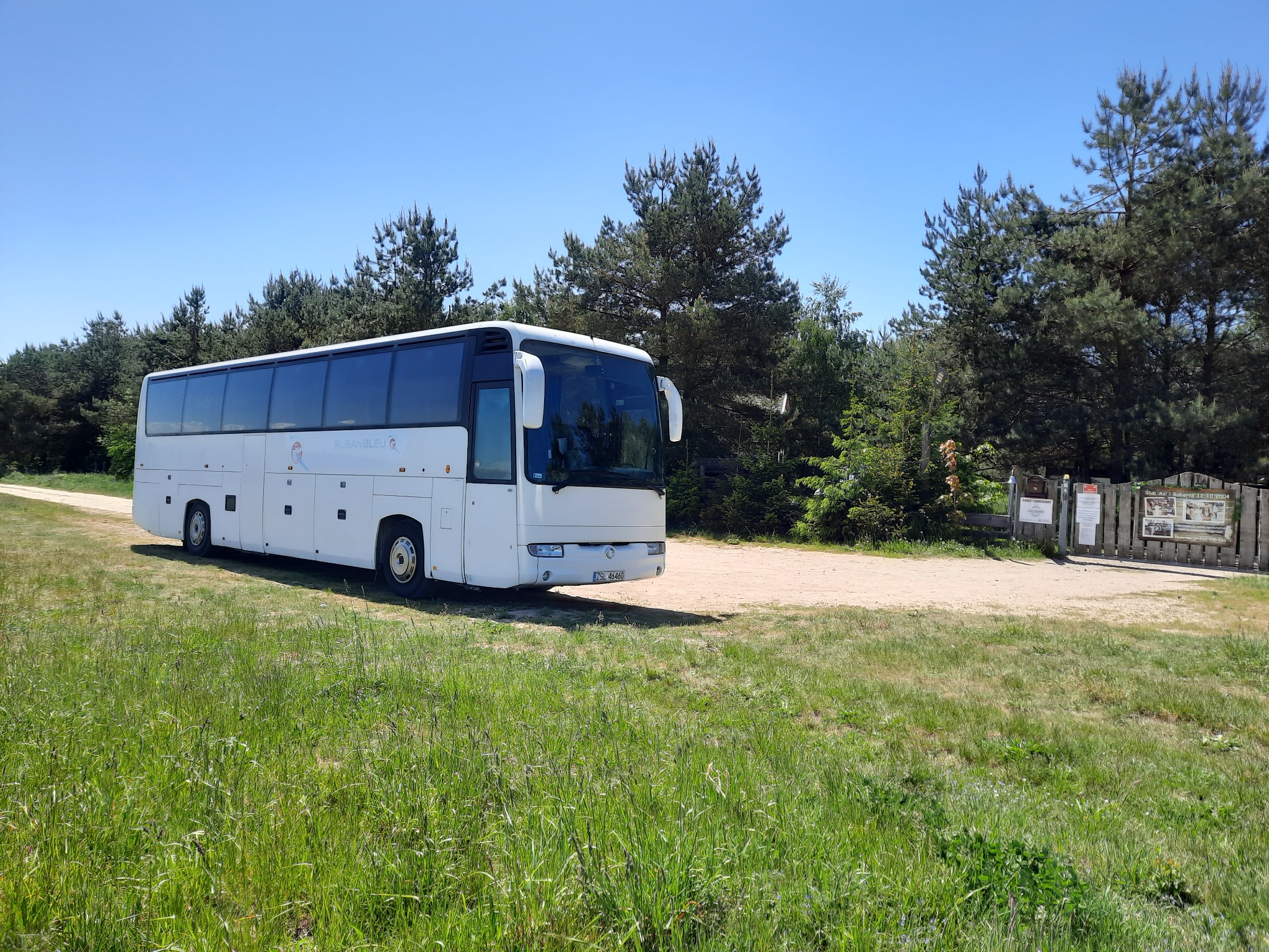 Biały autokar turystyczny zaparkowany na trawiastym terenie w pobliżu lasu, widoczny znak firmowy 'Ruban Bleu' na boku pojazdu.