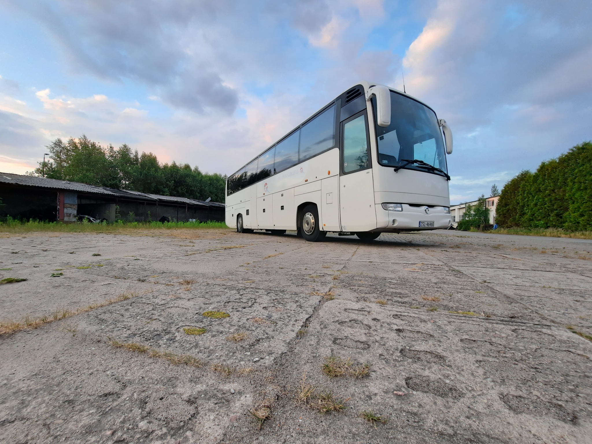 Biały autokar turystyczny zaparkowany na betonowym placu z mchem, na tle zachmurzonego nieba i zarośniętych budynków.