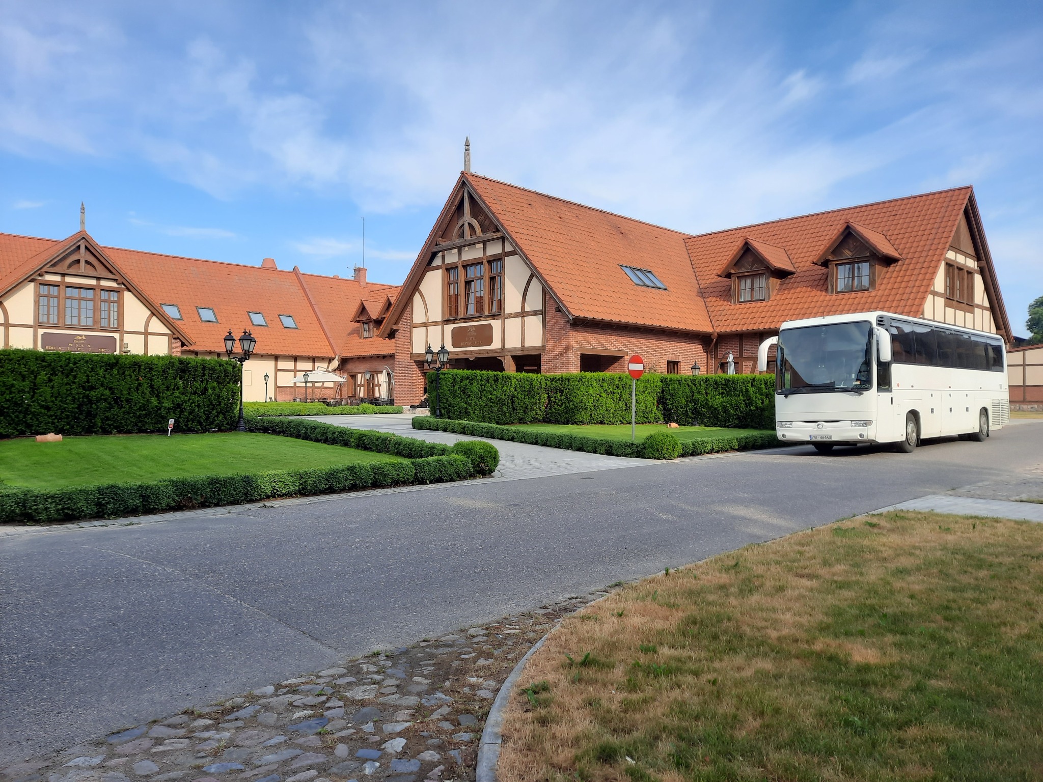 Biały autokar turystyczny zaparkowany przed budynkiem hotelowym z czerwoną dachówką i zadbanym trawnikiem.