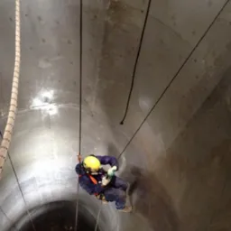 Pracownik w kasku, na linach, wewnątrz betonowego komina. Prace wysokościowe z użyciem specjalistycznego sprzętu. Widok z dołu, oświetlenie punktowe.