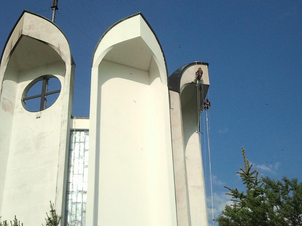 Dwóch alpinistów na linach, pracujących na wysokości na białej elewacji kościoła, widok z dołu, błękitne niebo w tle, fragment drzewa w prawym dolnym rogu.