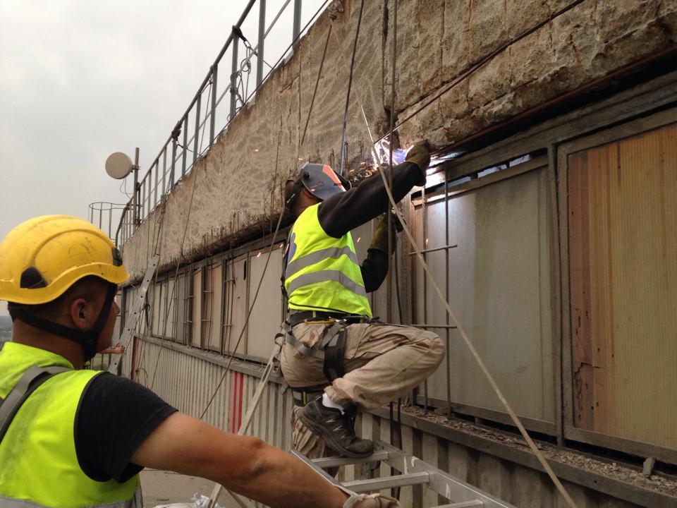 Spawanie wzmocnienia elewacji budynku przez pracownika w uprzęży, z drabiny. Widoczny drugi pracownik w żółtym kasku i kamizelce odblaskowej. Praca na wysokości.