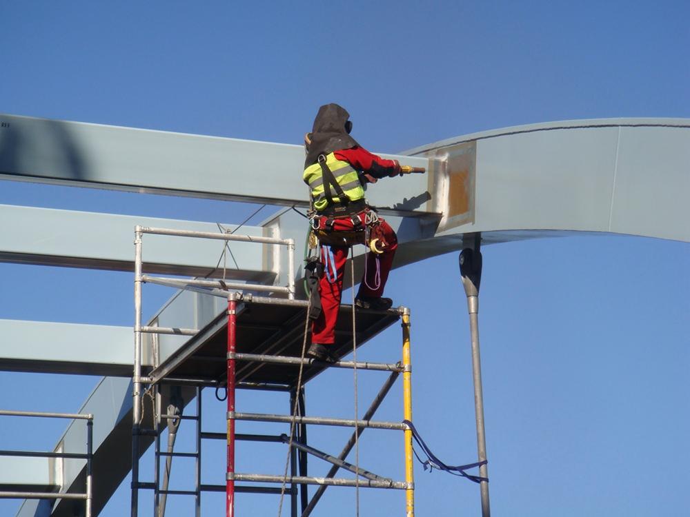 Pracownik w kombinezonie ochronnym na rusztowaniu szlifuje metalową konstrukcję na wysokości, przygotowując powierzchnię pod malowanie. Błękitne niebo w tle.