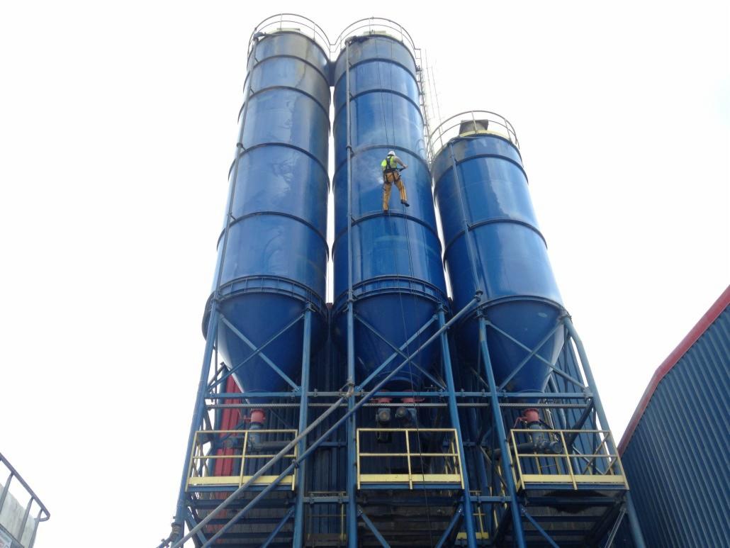 Pracownik w uprzęży na wysokości maluje błękitny silos. Silosy na stalowej konstrukcji, ujęcie z dołu, jasne niebo w tle. Praca na wysokościach.