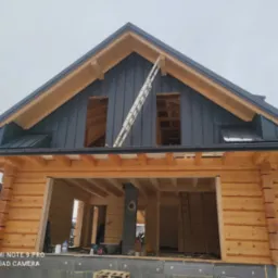 Surowa konstrukcja drewnianego domu z zamontowaną ciemną blachą na dachu i opartą drabiną o szczyt. Widoczne otwory okienne i drzwiowe, w tle zabudowa jednorodzinna.