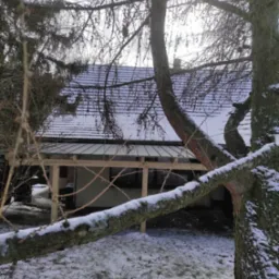 Zimowy krajobraz z widokiem na częściowo zadaszony taras przy domu, konstrukcja drewniana w surowym stanie, śnieg na dachu i gałęziach drzew.