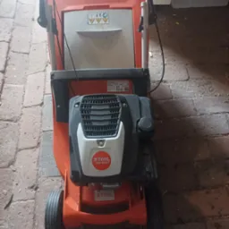Pomarańczowa kosiarka spalinowa Stihl RM 443 T, widok z góry na tle ceglanej posadzki i fragmentu szafki.
