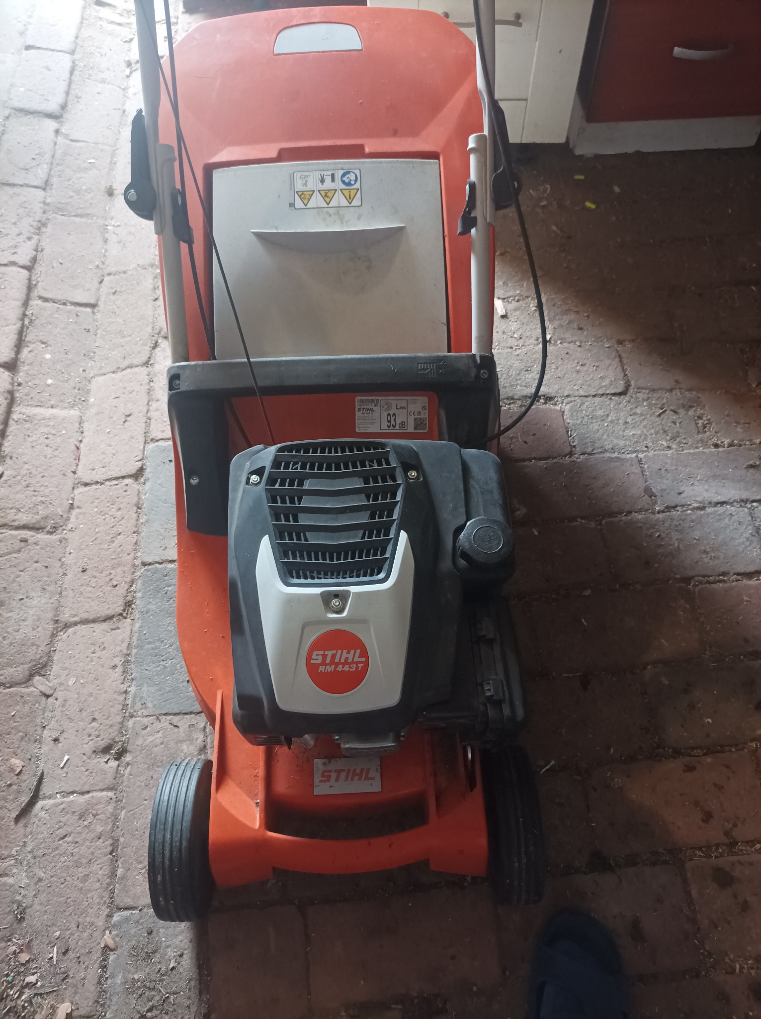 Pomarańczowa kosiarka spalinowa Stihl RM 443 T, widok z góry na tle ceglanej posadzki i fragmentu szafki.