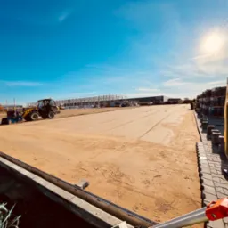 Żółta ładowarka kołowa na wyrównanym, piaszczystym terenie budowy z widocznymi paletami kostki brukowej i metalową konstrukcją budynku w tle, oświetlone jasnym słońcem.