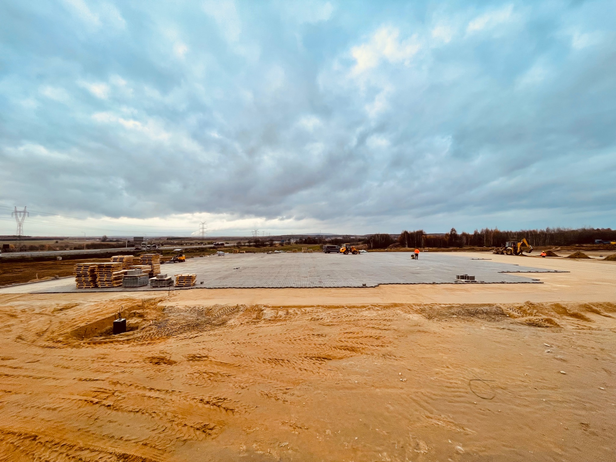 Szeroki kadr na plac budowy z częściowo ułożoną kostką brukową, paletami z kostką i piaskiem na pierwszym planie, oraz pracującymi maszynami budowlanymi w tle pod pochmurnym niebem.