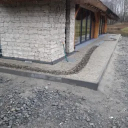 Wykończona elewacja budynku z kamienia z widocznym fragmentem dachu krytego strzechą, obok ułożona kostka brukowa i ozdobny pas z drobnych kamieni, w tle zbiornik wodny i drzewa.